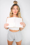 Blonde girl models the Namaste Home Loungewear Collection set for Scarlette The Label, an online fashion boutique for women. The set includes a soft white crop top and gray loungewear shorts. Sold separately.