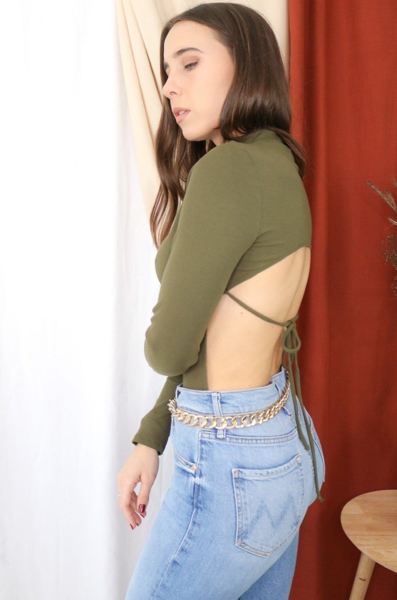 Brunette models long sleeve openback tie bodysuit in color olive for Scarlette The Label, an online fashion boutique for women. Paired with lightwash denim jeans and a gold chain belt. 