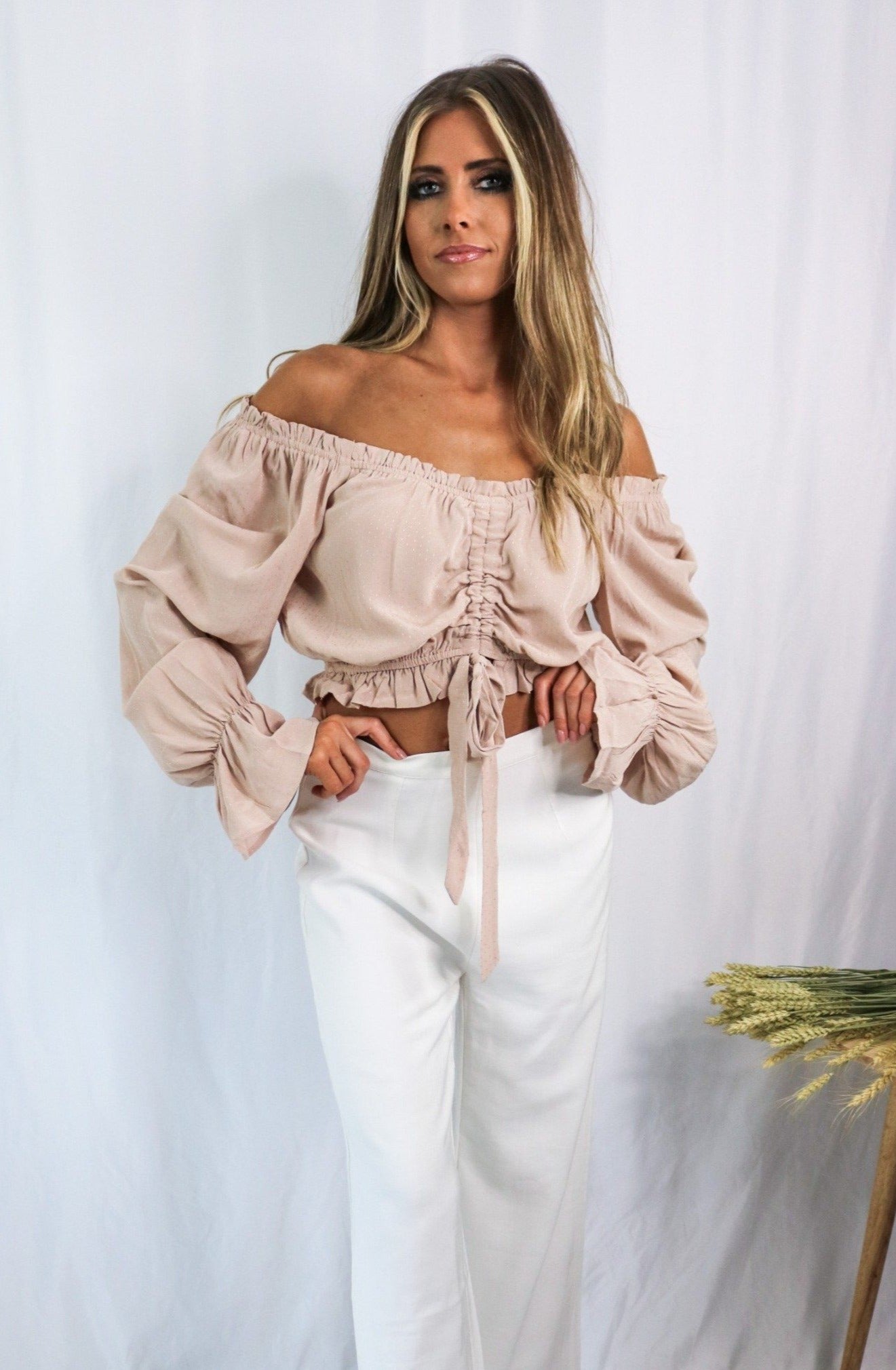 Blonde girl models an off the shoulder frilled blouse in color blush for Scarlette The Label, an online fashion boutique for women. Paired with white, wide-leg pants.