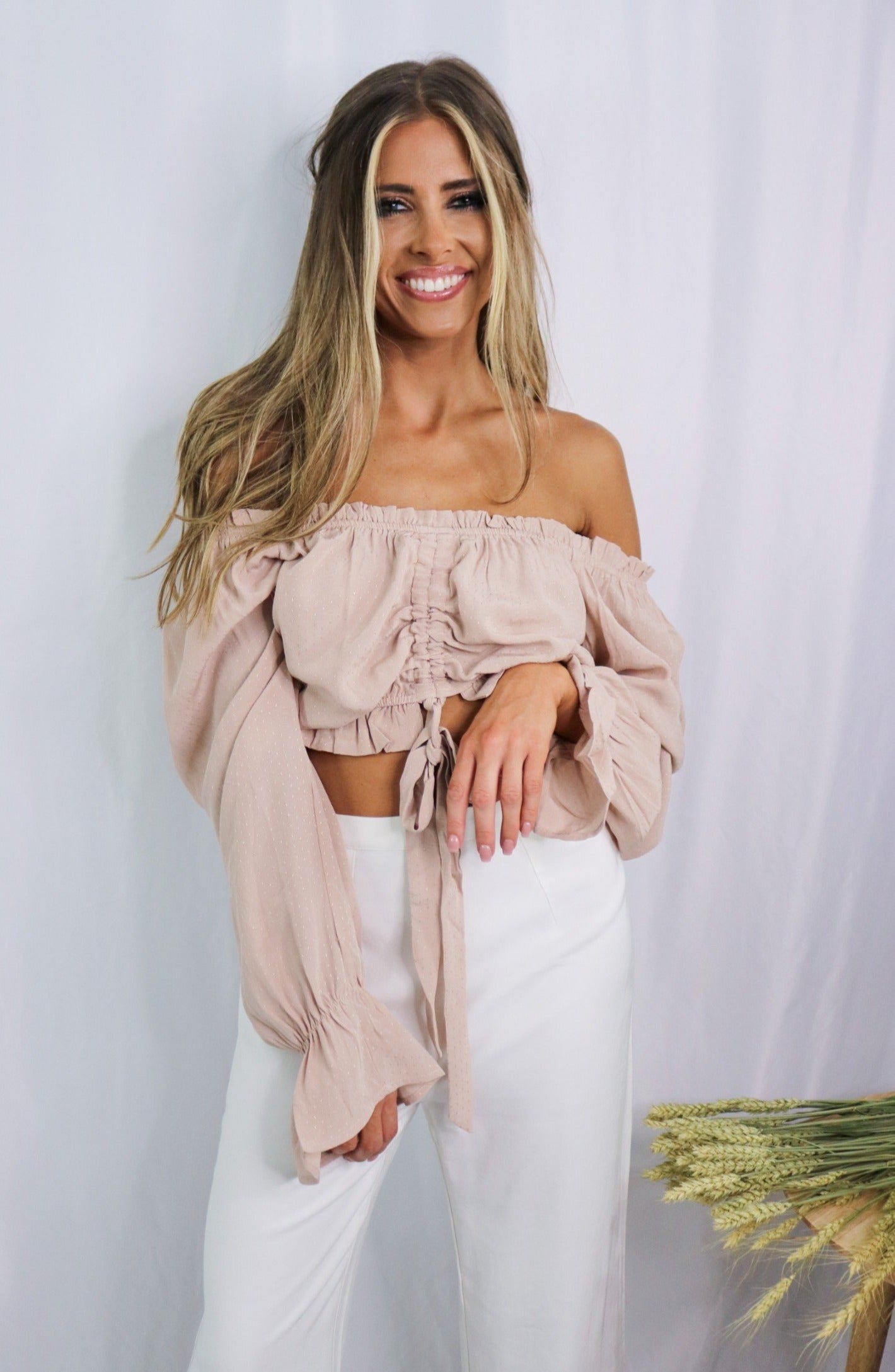 Blonde girl models an off the shoulder frilled blouse in color blush for Scarlette The Label, an online fashion boutique for women. Paired with white, wide-leg pants.