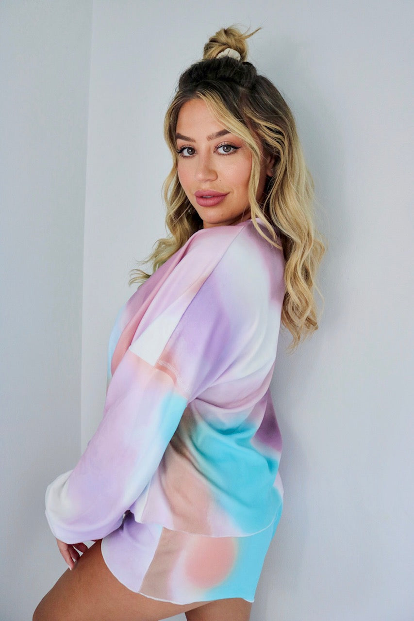 Blonde girl models a tie dye loungewear short set for Scarlette The Label, an online fashion boutique for women. The loungewear set is light blue, light purple and light pink and is a tie dye pattern. The matching tie-dye loungewear set includes a long sleeve shirt and shorts.