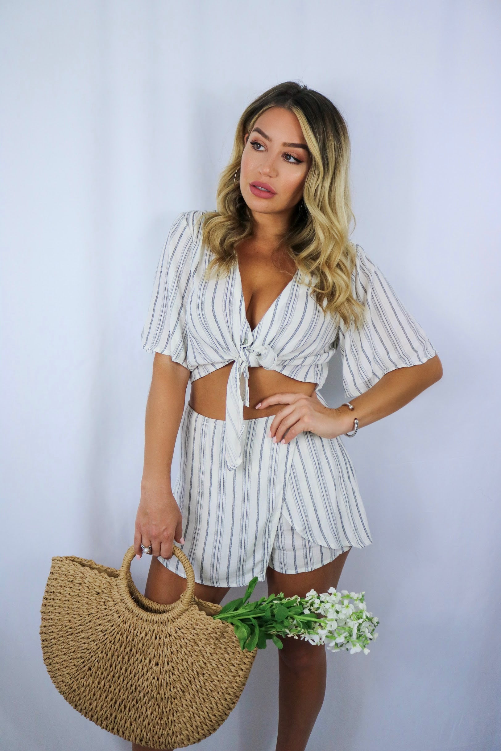 Blonde girl models a matching set for Scarlette The Label, an online fashion boutique for women. The set is a matching short set in white and charcoal stripes. Includes a short sleeve tie top and a skort. Paired with large straw bag.