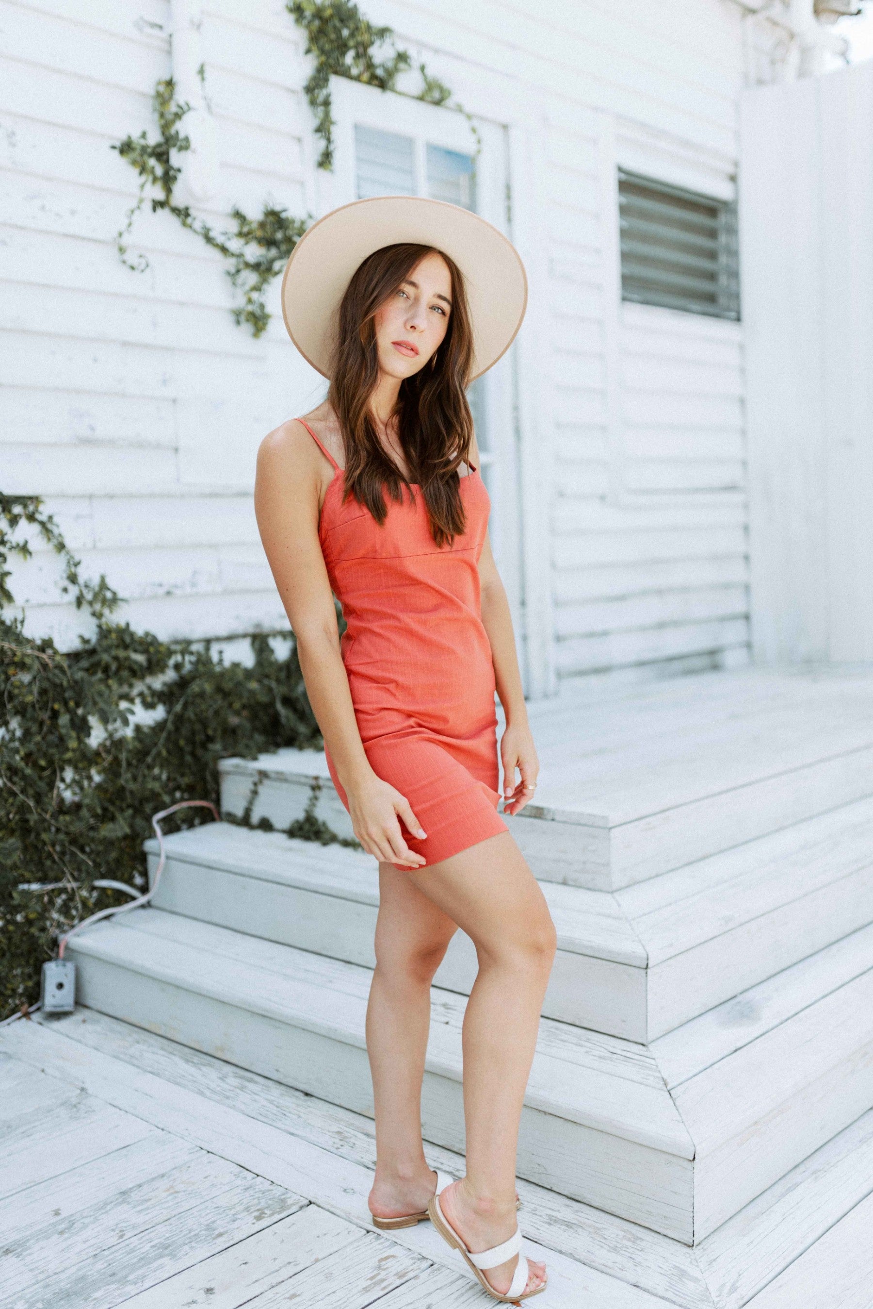 Brunette girl models a solid orange spaghetti strap mini dress for Scarlette The Label, an online fashion boutique for women. The orange mini dress is paired with a wide-brimmed rancher hat.