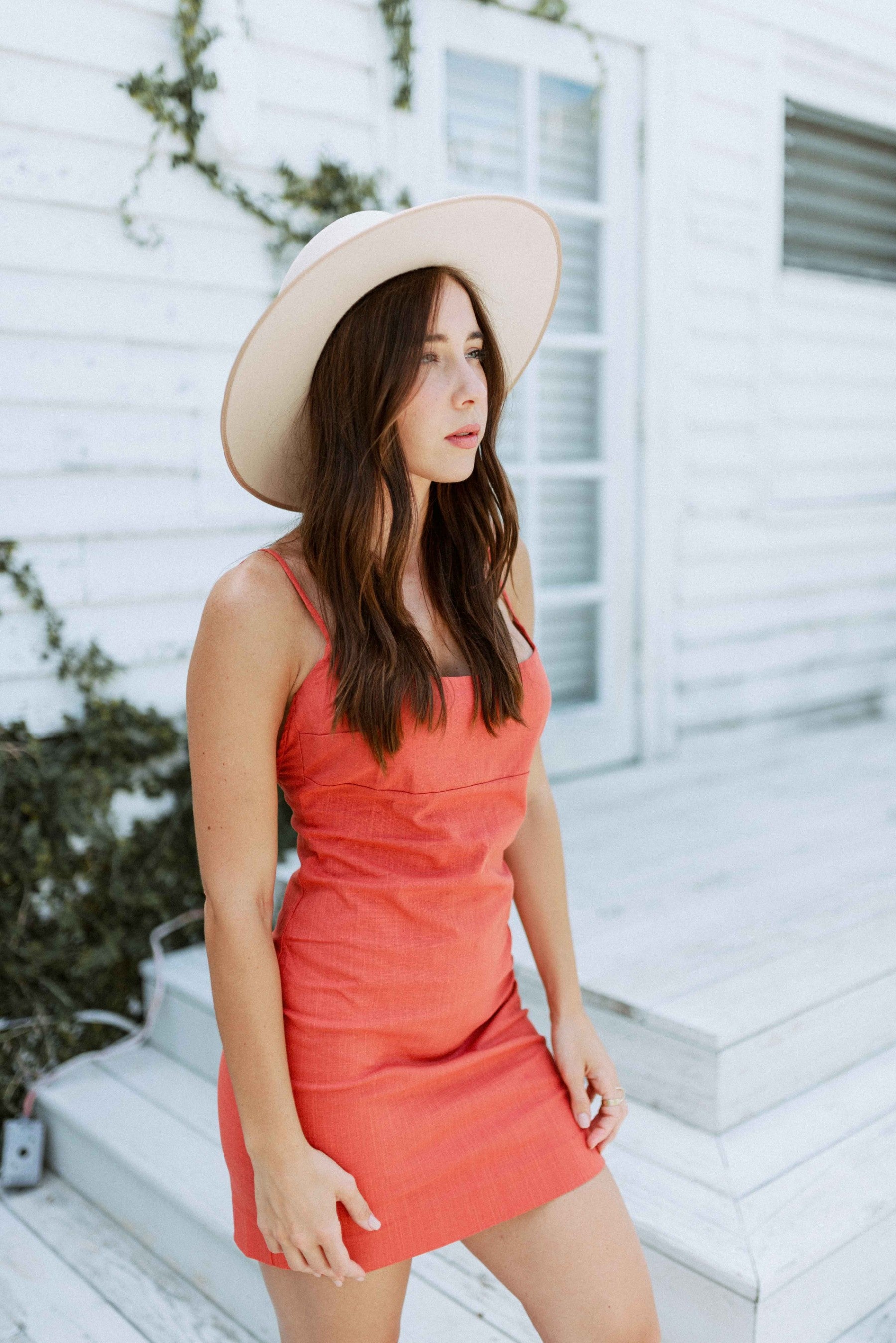 Brunette girl models a solid orange spaghetti strap mini dress for Scarlette The Label, an online fashion boutique for women. The orange mini dress is paired with a wide-brimmed rancher hat.