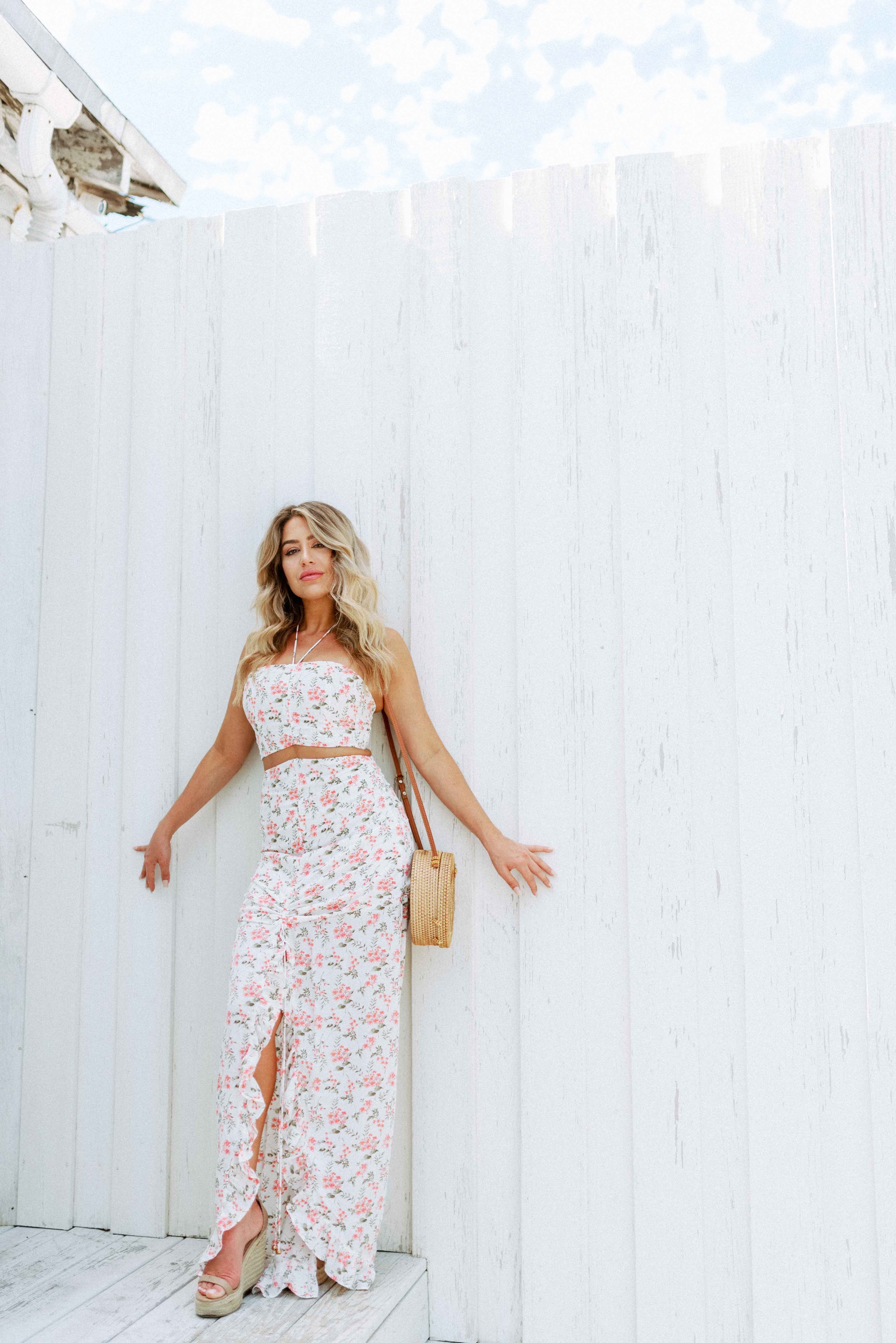 Blonde girl models a floral maxi skirt matching set for Scarlette The Label, an online fashion boutique for women.  The matching skirt set has a halter crop top and a long slitted maxi skirt. Sold separately. Paired with straw purse.