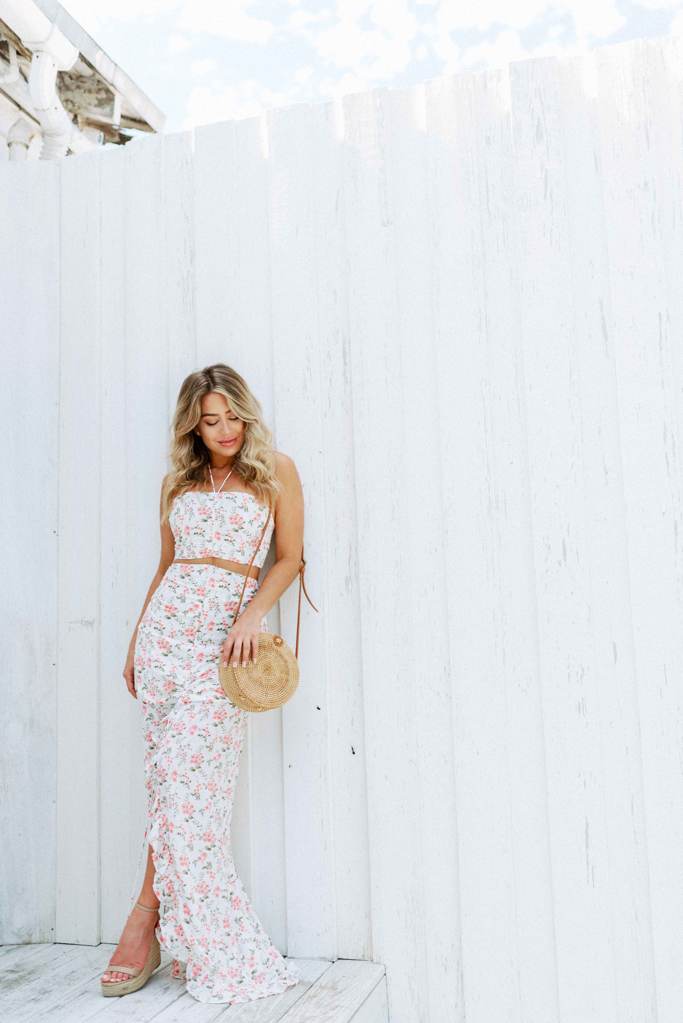 Blonde girl models a floral maxi skirt matching set for Scarlette The Label, an online fashion boutique for women.  The matching skirt set has a halter crop top and a long slitted maxi skirt. Sold separately. Paired with straw purse.